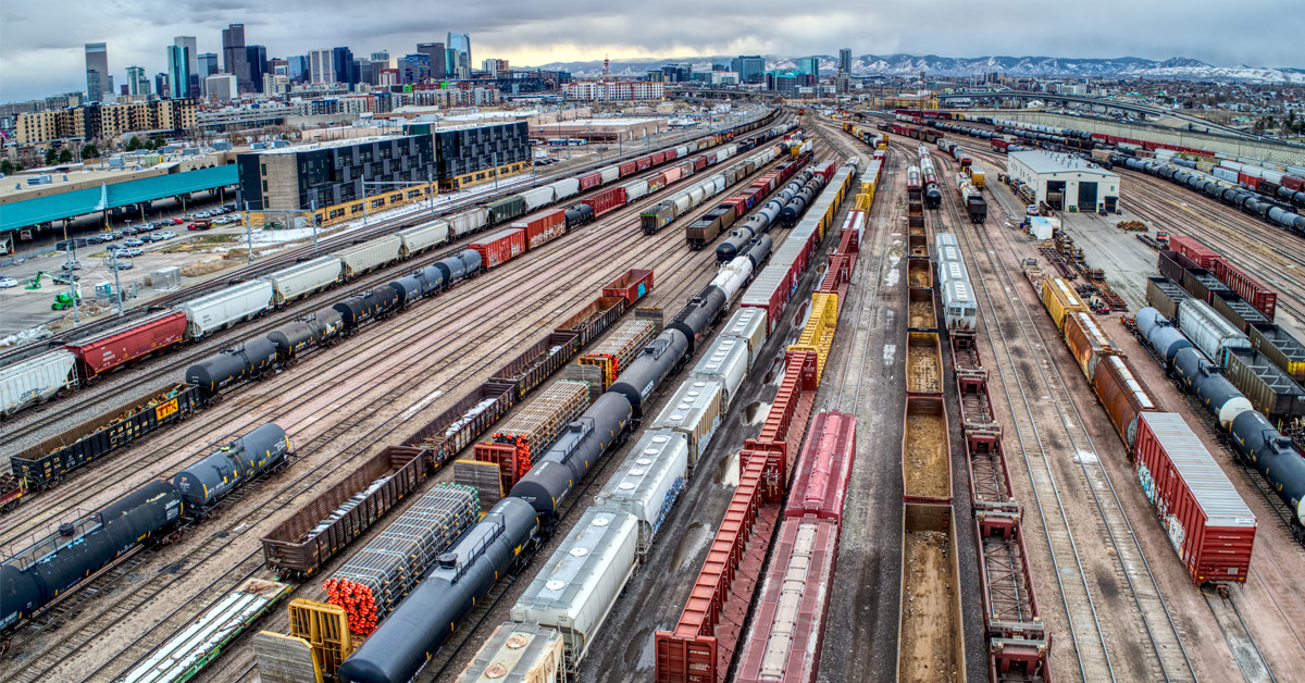 A rail yard with lines of colorful, varied railcars with a city skyline in the distance.