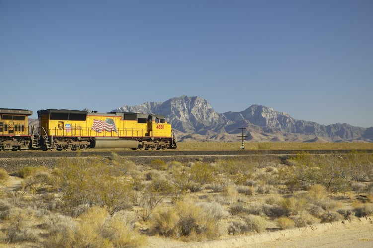 A yellow train with an american flag on the side travels through a desert in front of mountains.